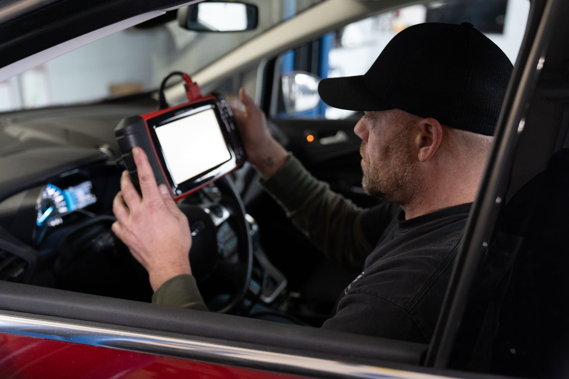 Mechanic in a car using a diagnostic tool with a blank screen.