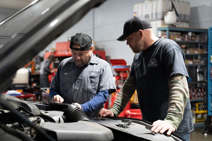 Two mechanics examine a car engine in a garage. One holds a tablet, both wearing caps.