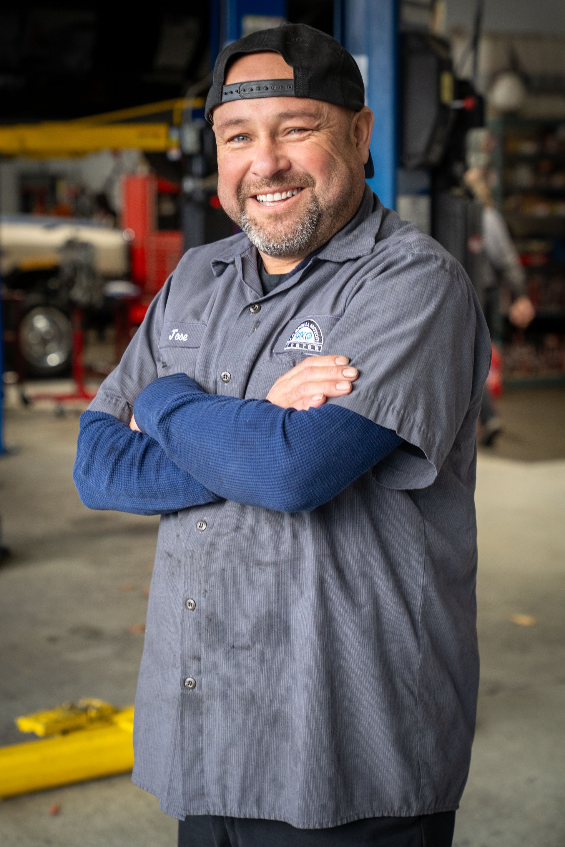 Smiling mechanic with arms crossed, in a shop wearing a cap, gray shirt, and blue sleeves.