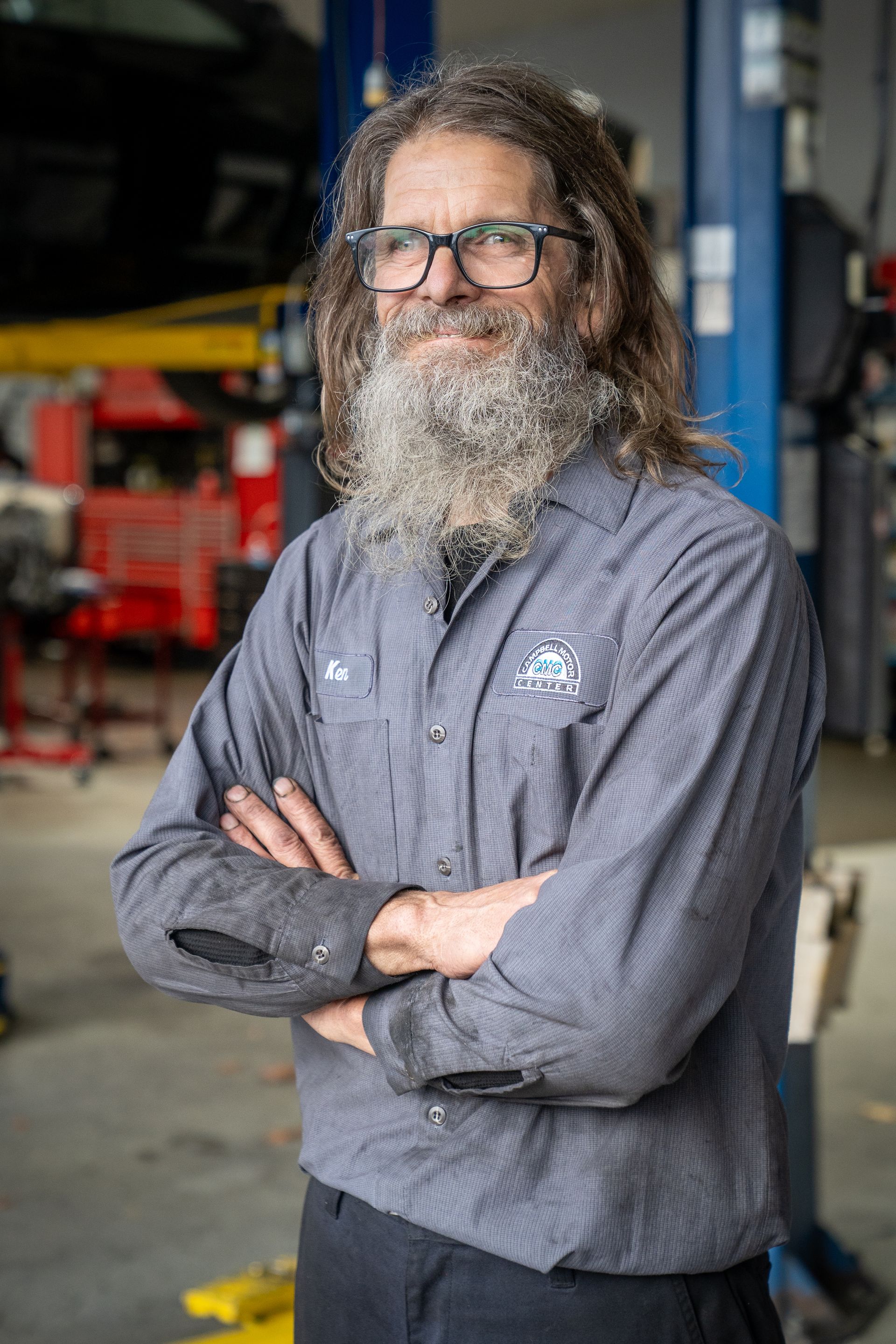 Mechanic with grey beard and glasses, arms crossed, in a repair shop.