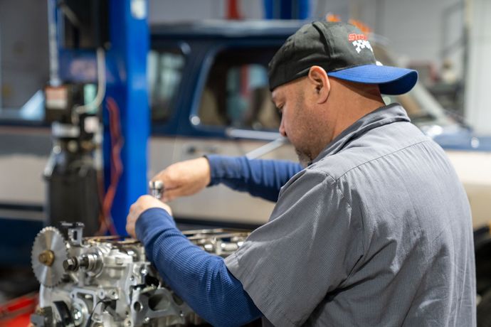 Mechanic working on an engine in a garage; wearing a hat, blue shirt, and using a wrench.