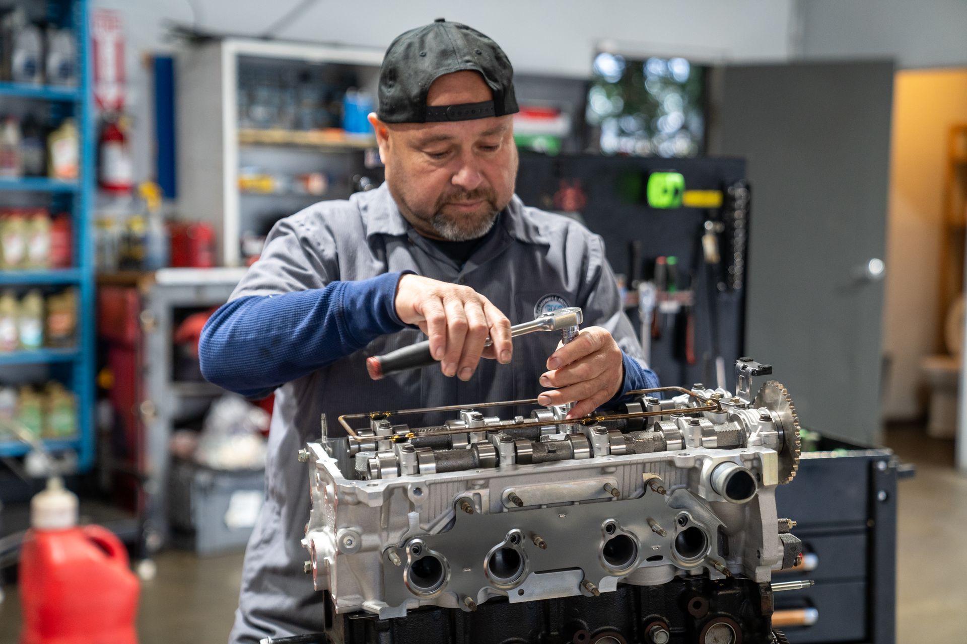 Mechanic in a gray work shirt and black cap uses a wrench on an engine in a garage.