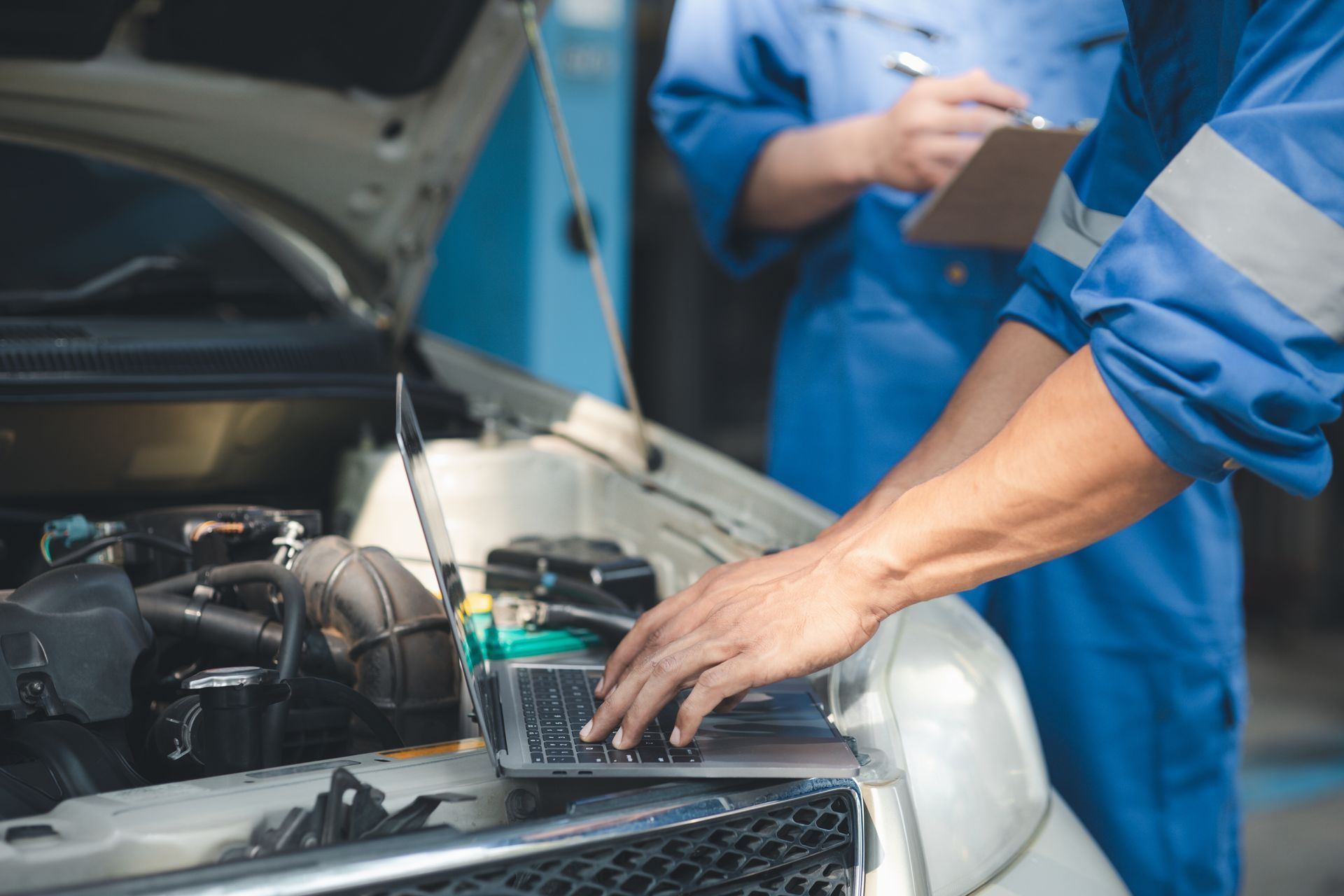 Mechanic using a laptop to diagnose a car engine; another person in overalls watches.