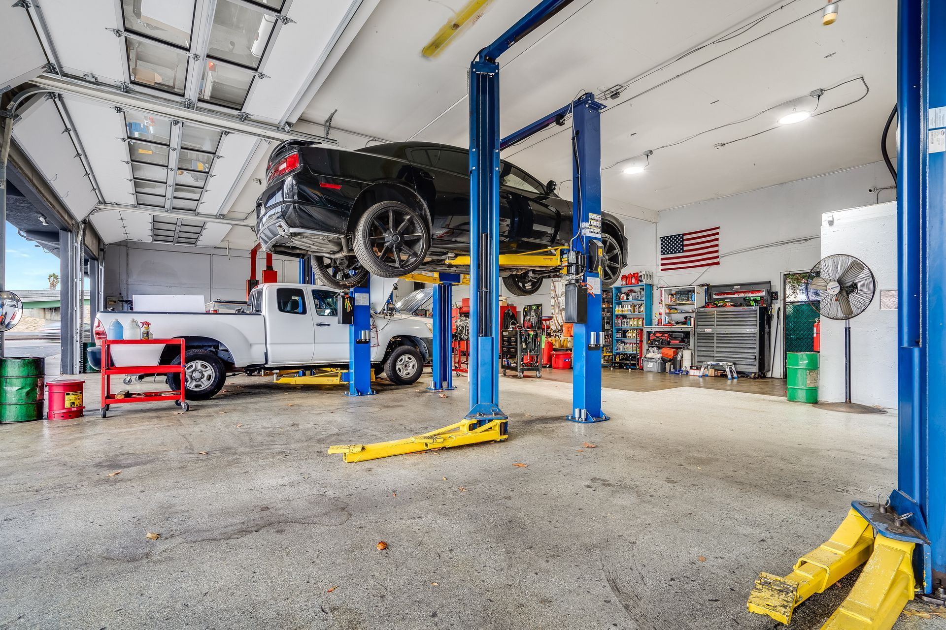Inside a car repair shop, a black car is lifted, a white truck is parked, and tools are visible.