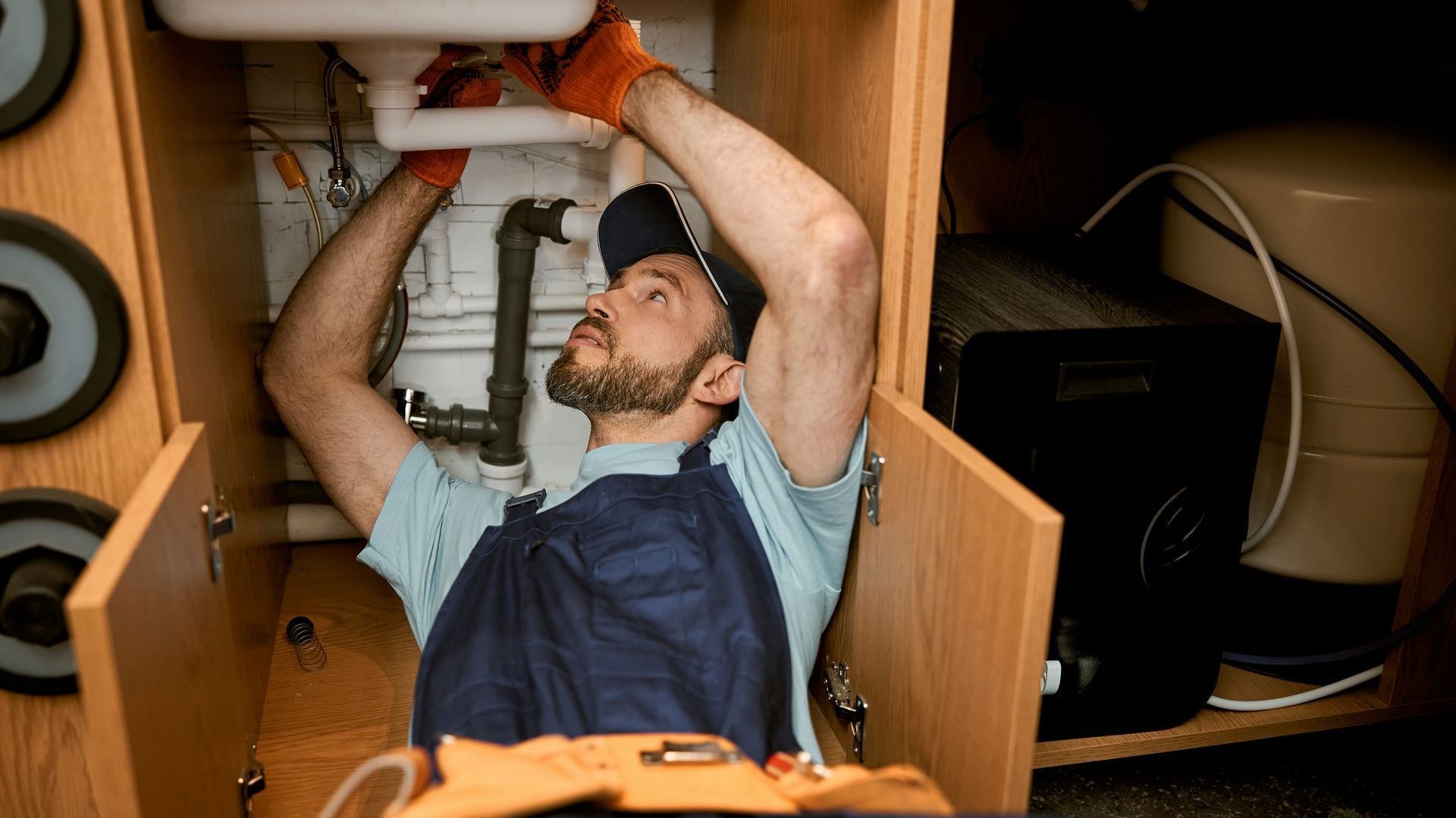 A man is fixing a sink under a cabinet in a kitchen.