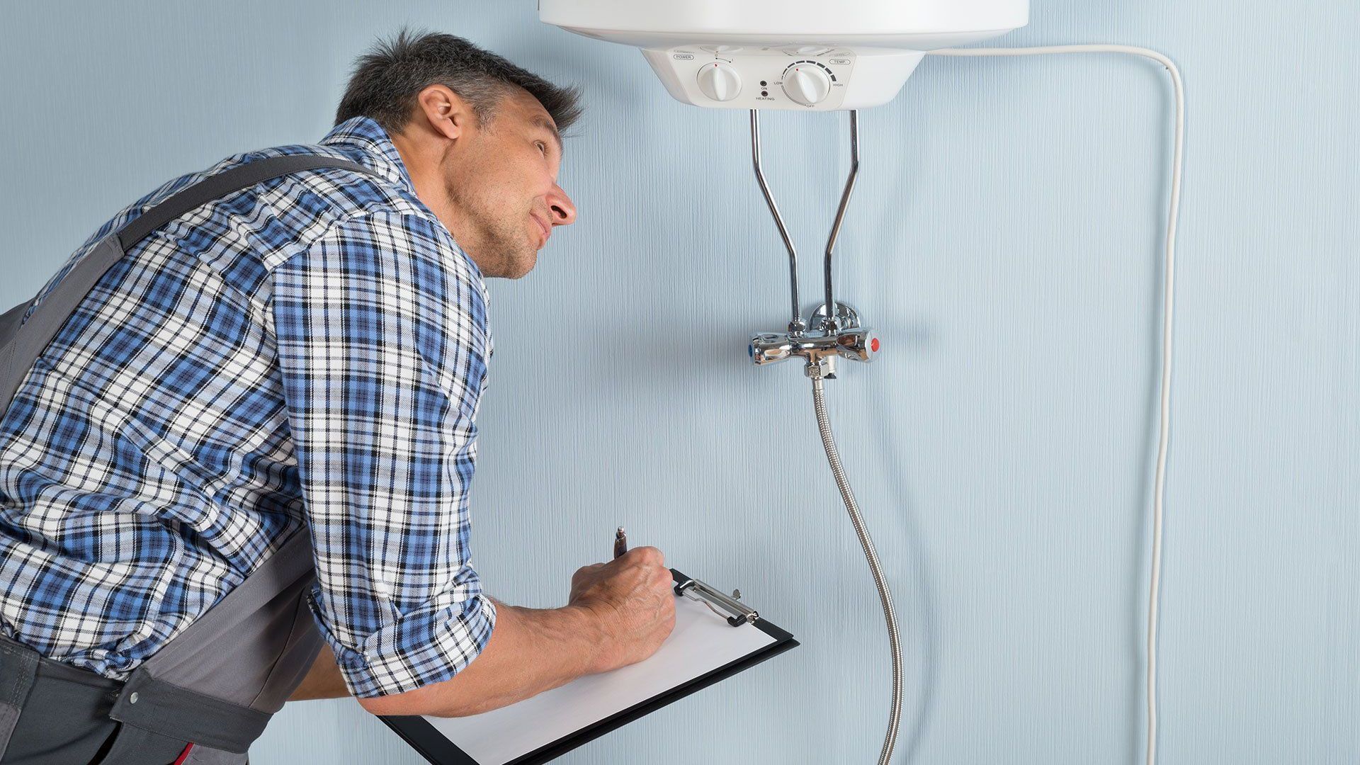 A man is looking at a water heater while holding a clipboard.
