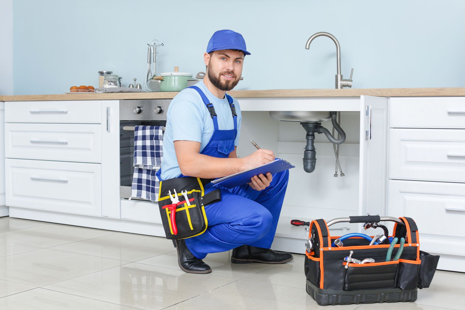 A plumber is kneeling under a sink in a kitchen.