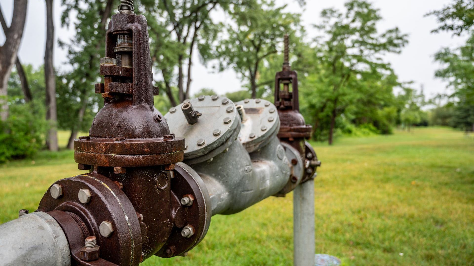 A close up of a rusty pipe in a field with trees in the background.