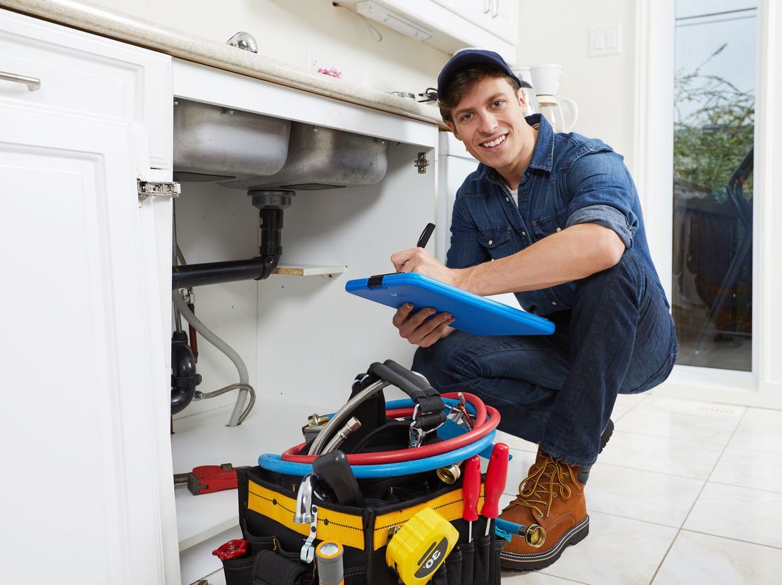 A plumber is kneeling under a sink in a kitchen and writing on a clipboard.