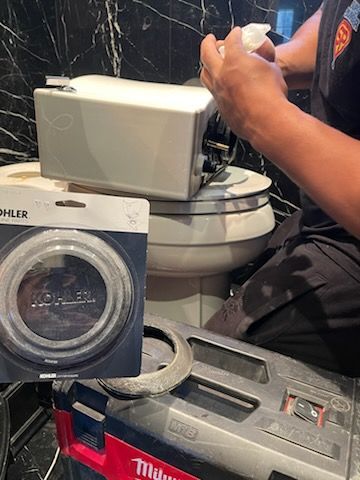 Person repairing a toilet, holding a part, with a new Kohler seal next to the toilet.