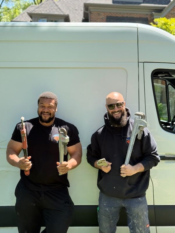 Two men holding wrenches, standing in front of a white van. One wears black, the other, a black hoodie.