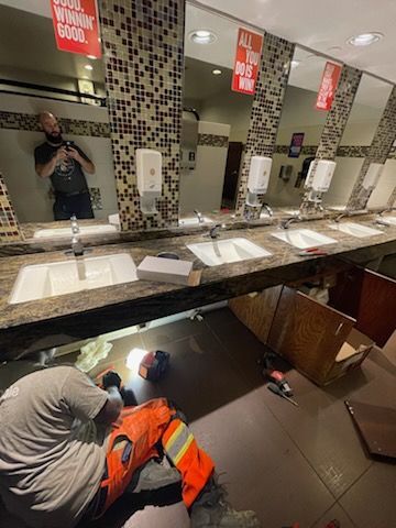 Bathroom interior: person photographing mirror, two workers repairing cabinets below sinks, white sinks, brown counters.