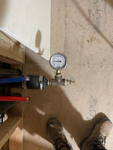 Plumbing with pressure gauge: blue and red pipes, black fitting, gauge at 50 psi. Gray concrete floor, work boots visible.