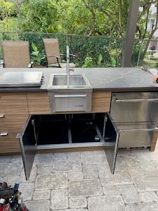 Outdoor kitchen with sink, grill, and open cabinet doors. Brown cabinets and stone countertop.