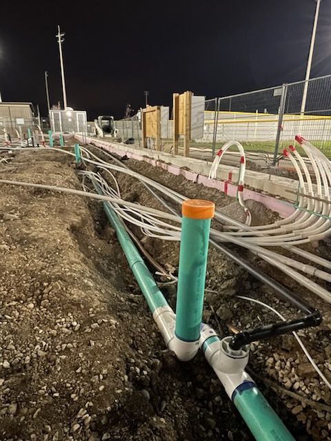 Construction site at night. Green pipes and white cables in a trench, orange capped pipe, materials in the background.
