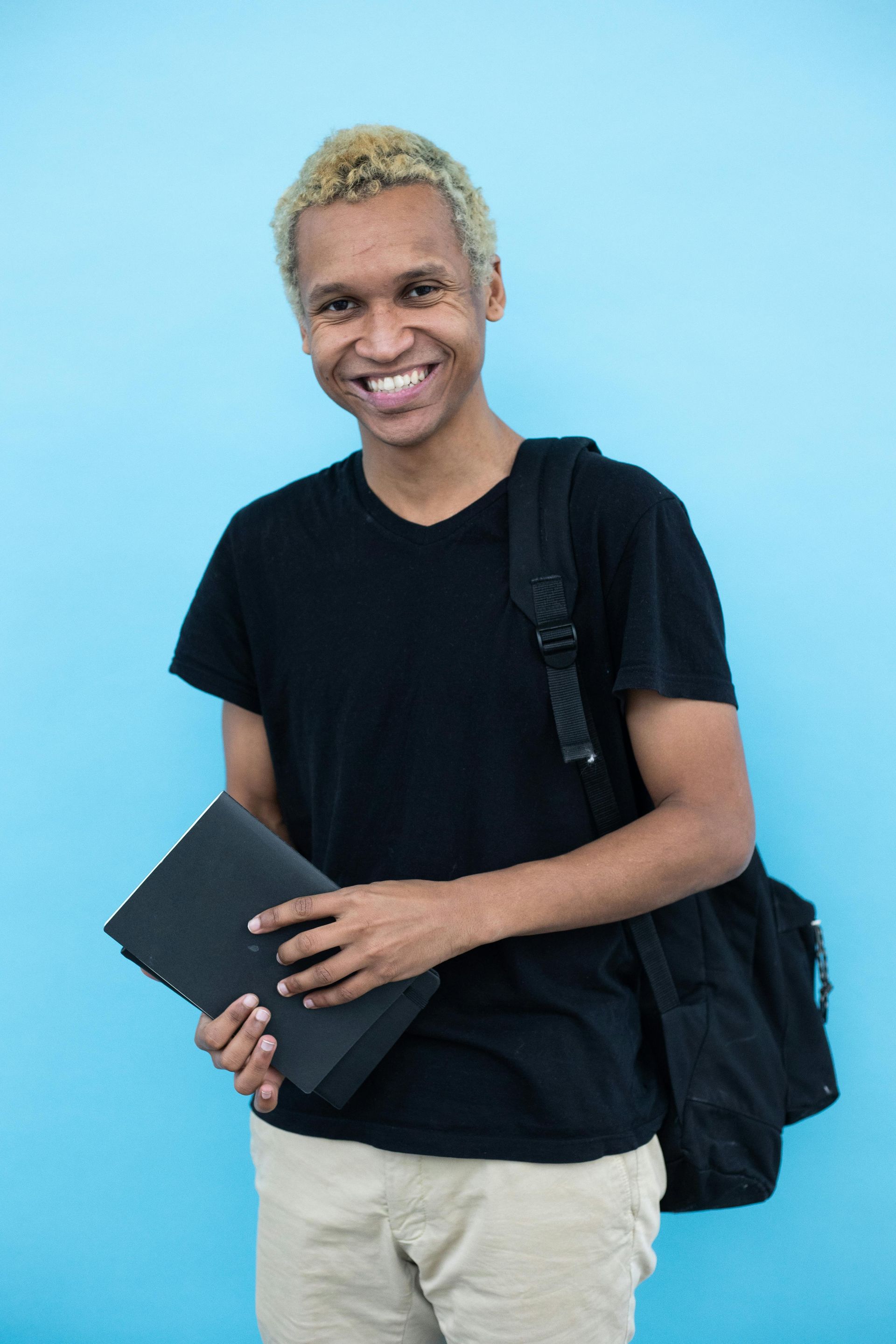 A young man is holding a laptop and smiling while standing in front of a blue wall.