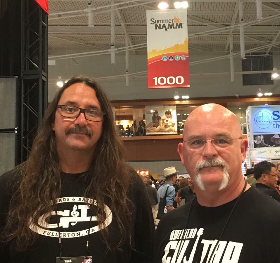Two men at a NAMM show. One has long hair, the other has a bald head. Both have mustaches and are looking at the camera.
