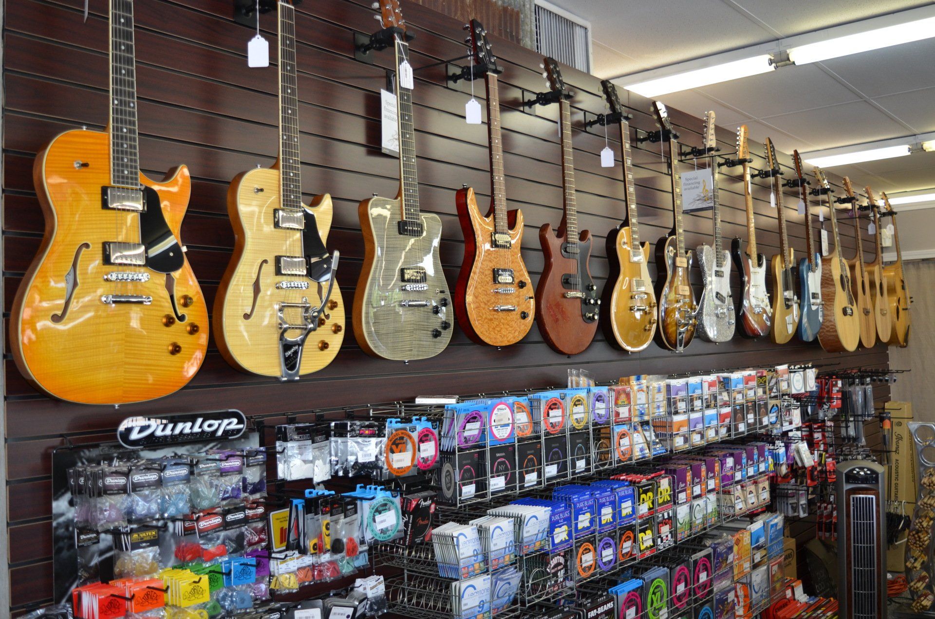 Guitars on display at a music store with guitar accessories below. Brown wall.