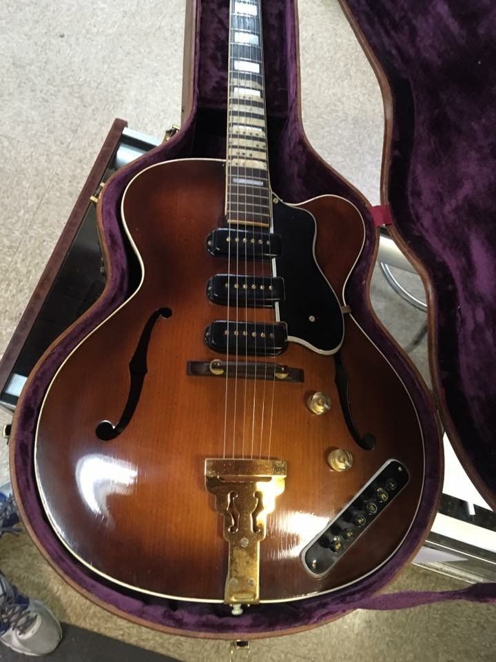 Brown hollow-body electric guitar in open case with purple lining, showing f-holes, three pickups, and gold hardware.