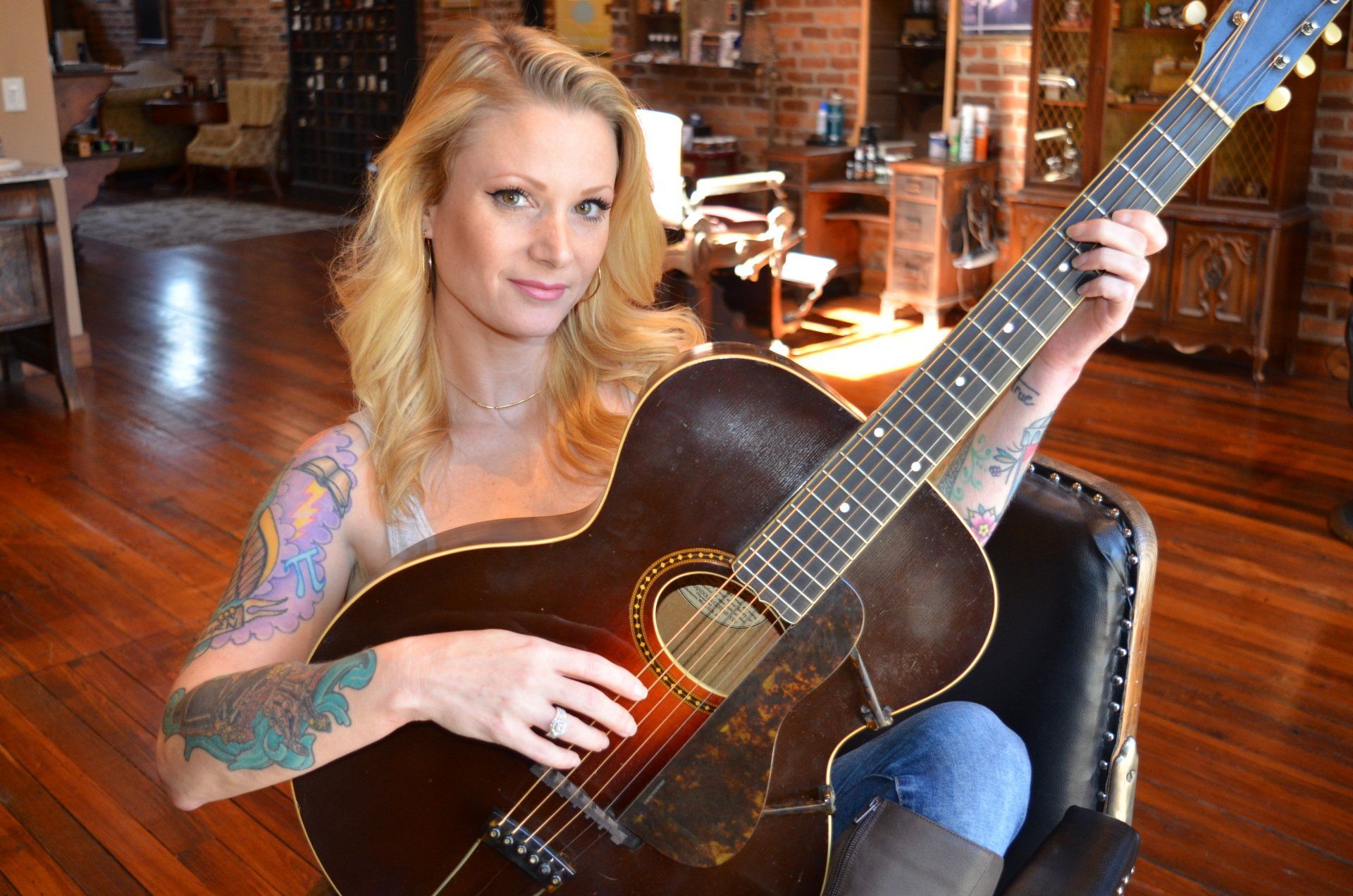 Woman with blonde hair, tattooed arms, playing an acoustic guitar. Sitting in a chair indoors.