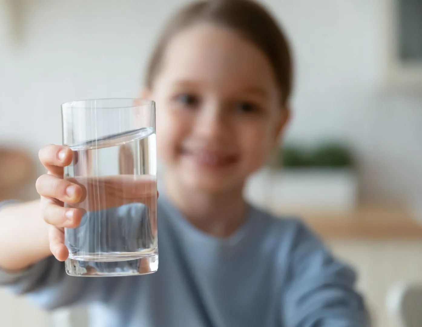 Un niño sostiene un vaso de agua transparente en una habitación luminosa y borrosa.