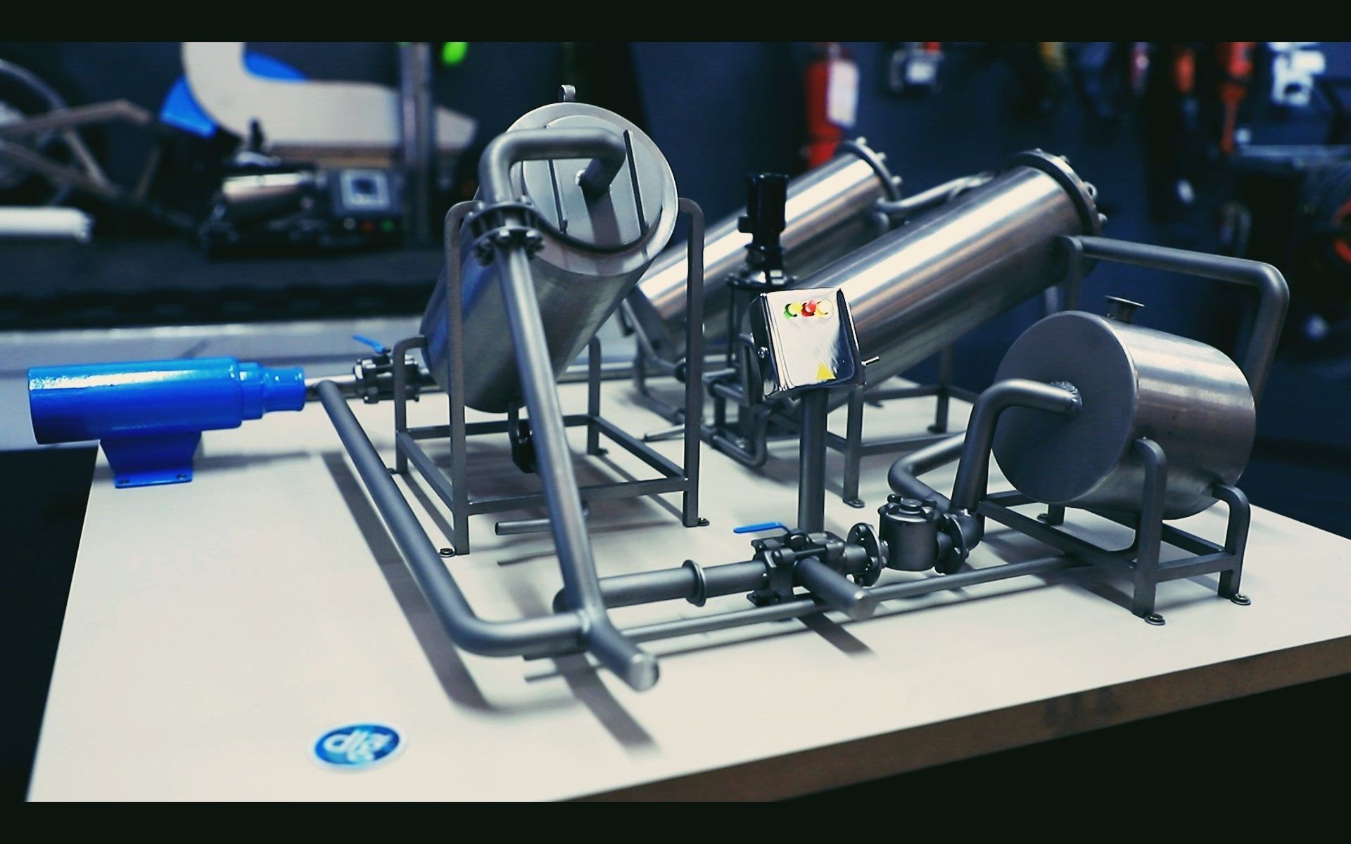 Prototipo de maquinaria industrial metálica sobre una mesa, con un rodillo cilíndrico, engranajes y una palanca azul.