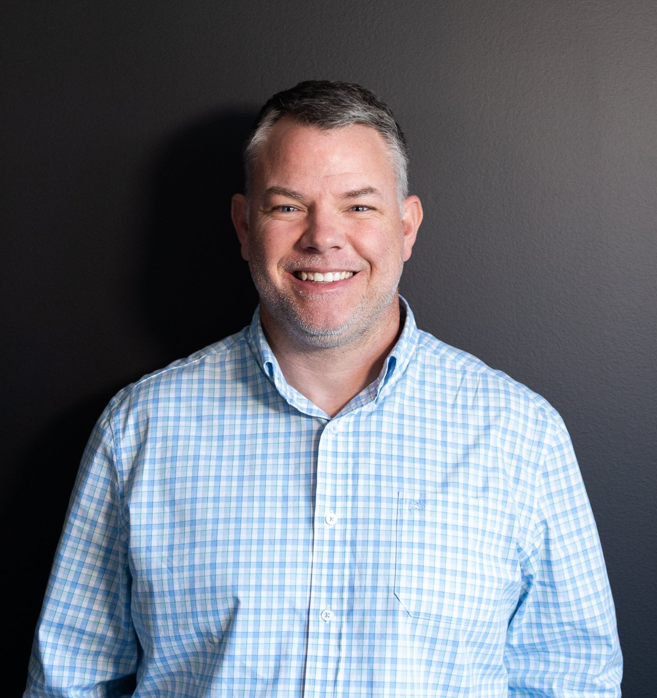A man in a blue plaid shirt is smiling in front of a black wall.