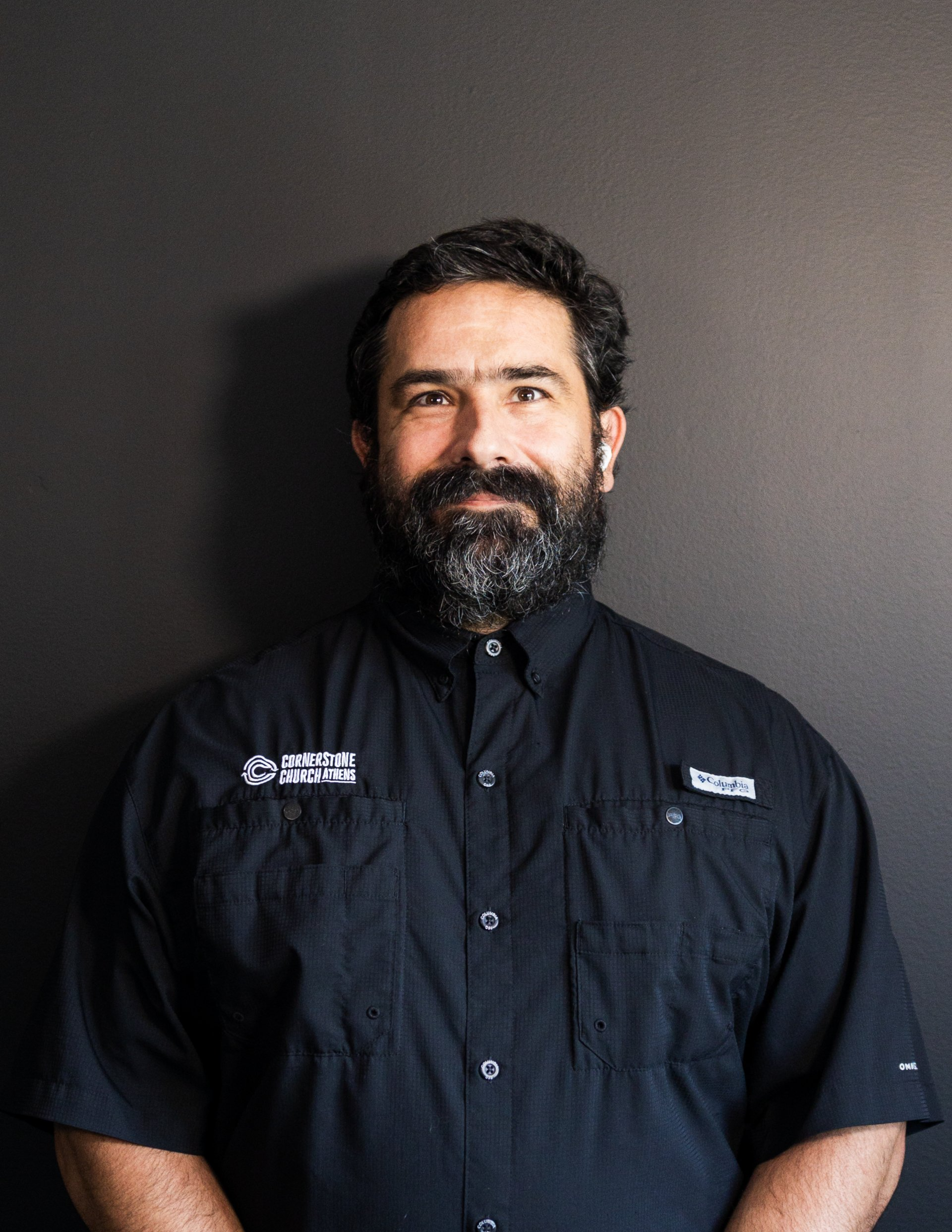 A man with a beard wearing a black shirt is standing in front of a black wall.