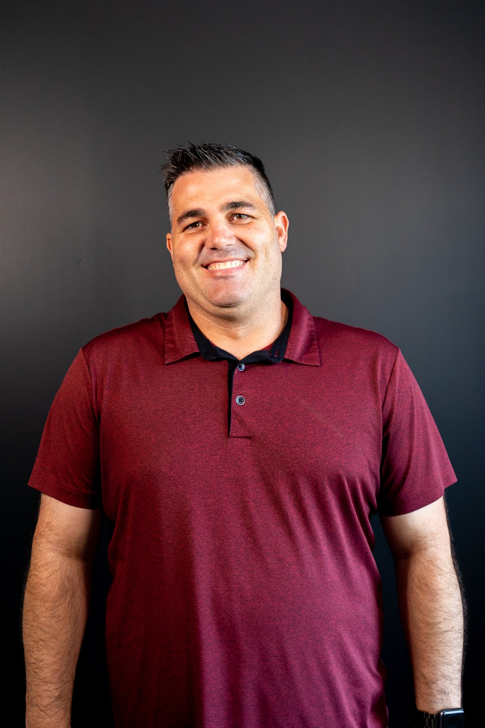 A man in a maroon polo shirt is smiling and standing in front of a black wall.