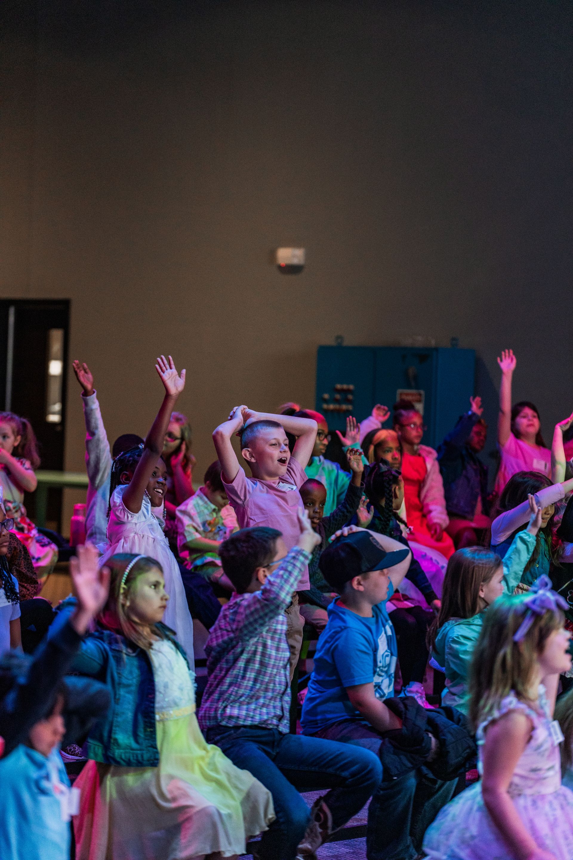 A group of children are sitting on the floor with their hands in the air.