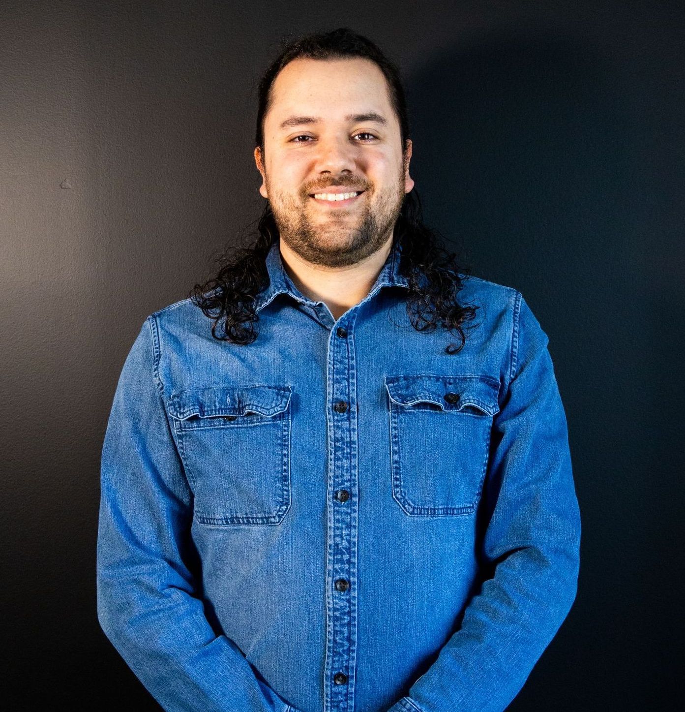 A man with long hair and a beard is wearing a blue denim shirt.