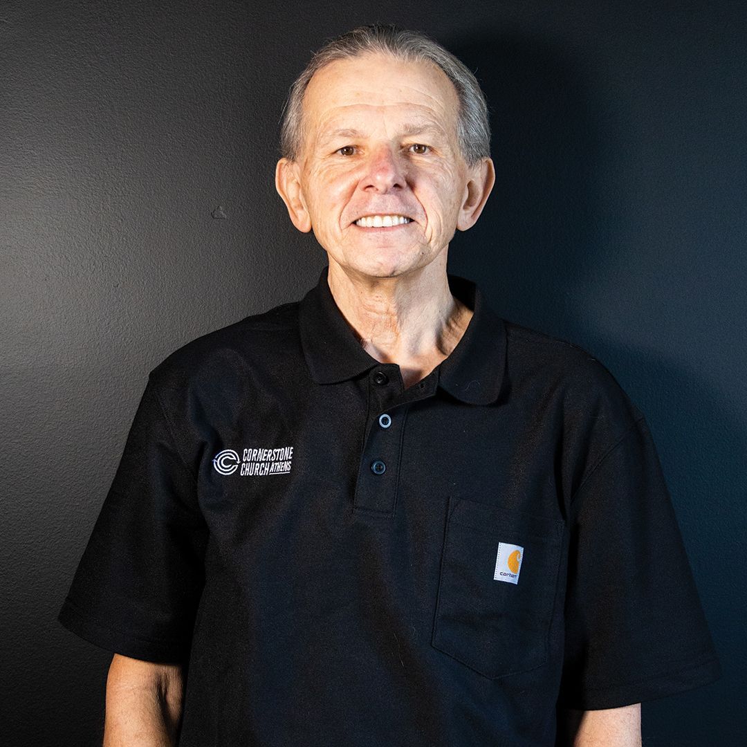 A man wearing a black polo shirt is standing in front of a black wall.