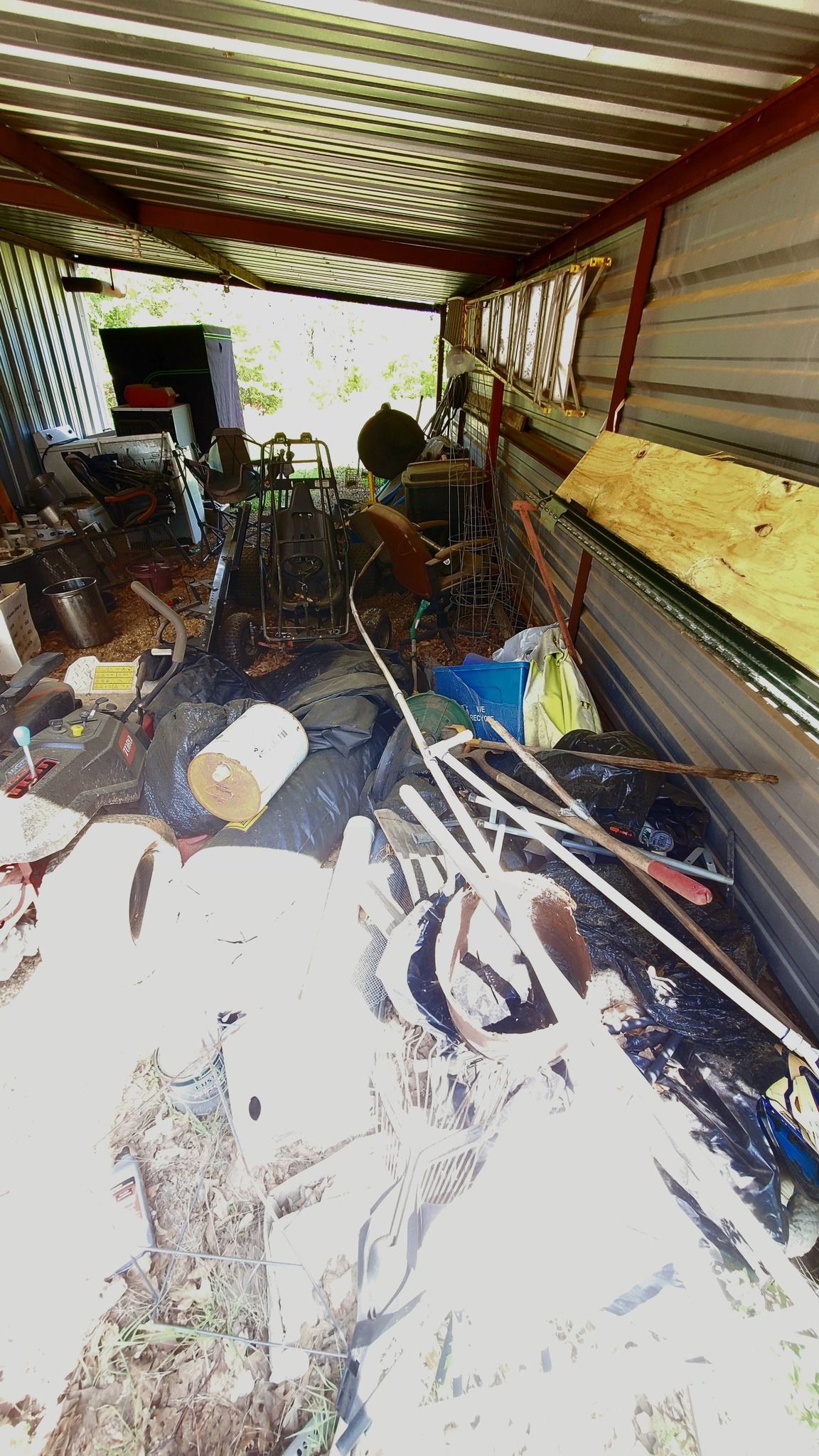 A cluttered, sunlit storage shed containing miscellaneous items, scrap metal, and tools under a corrugated metal roof.
