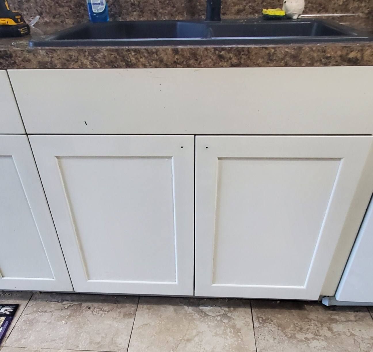 A kitchen sink with a dark basin set into a brown countertop above plain, white shaker-style base cabinets.