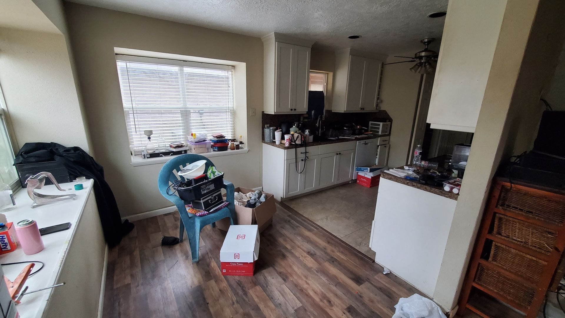 A cluttered dining area flows into a kitchen with white cabinets, wood-look flooring, and a window with white blinds.