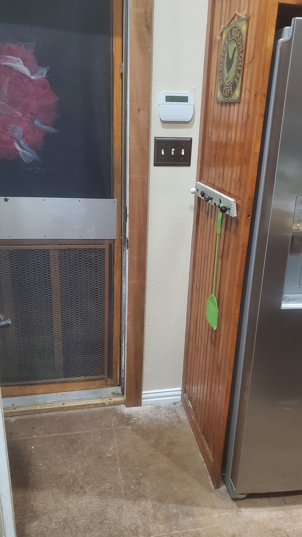 A wooden refrigerator panel stands near a doorway with a screen door and a wall-mounted security panel and light switch.
