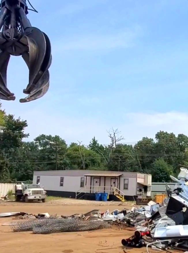 A metal claw hangs over a scrap yard with a truck and mobile home in the background.