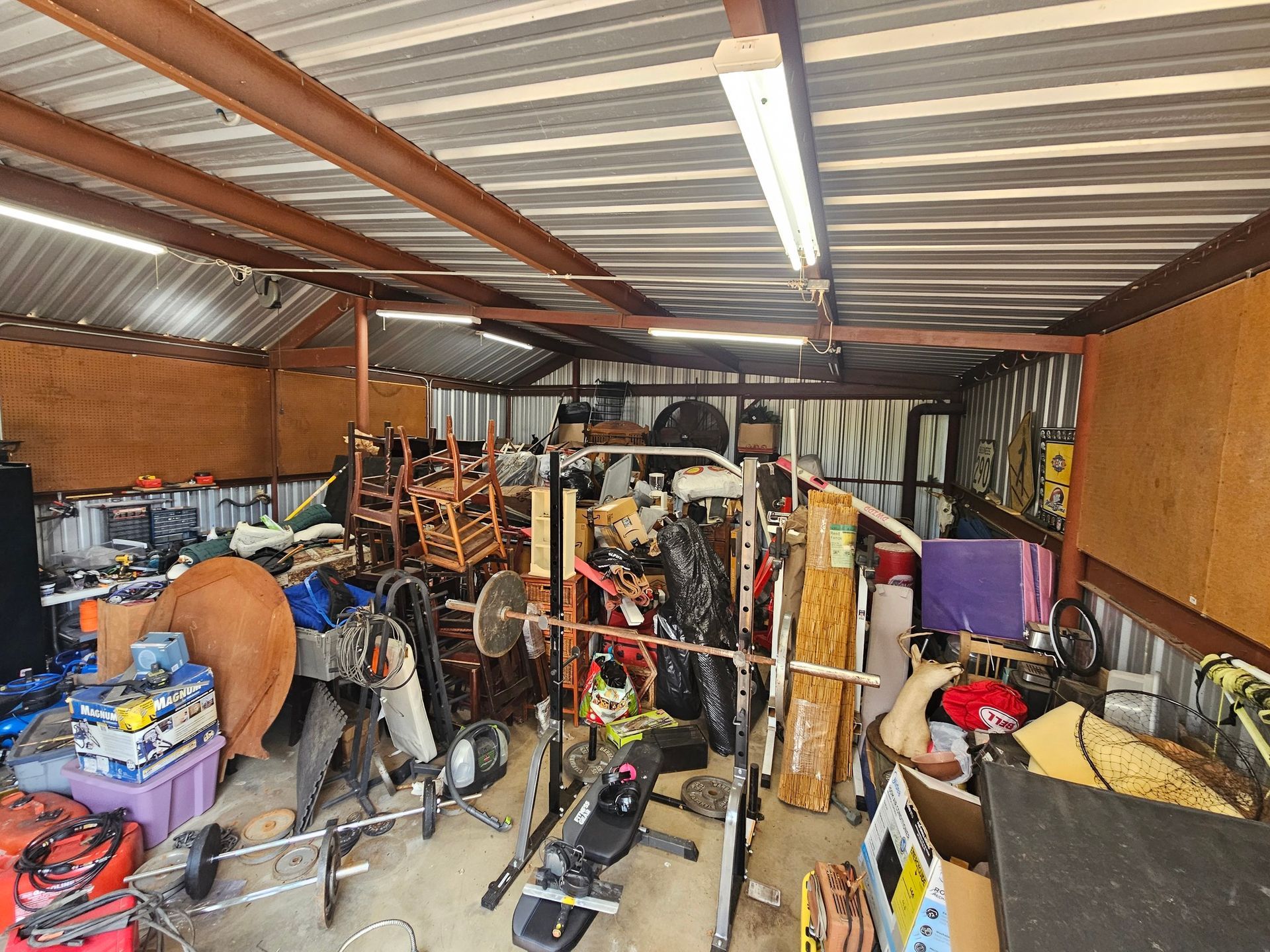 A cluttered garage with a gym rack, various tools, storage boxes, and household items scattered throughout the space.