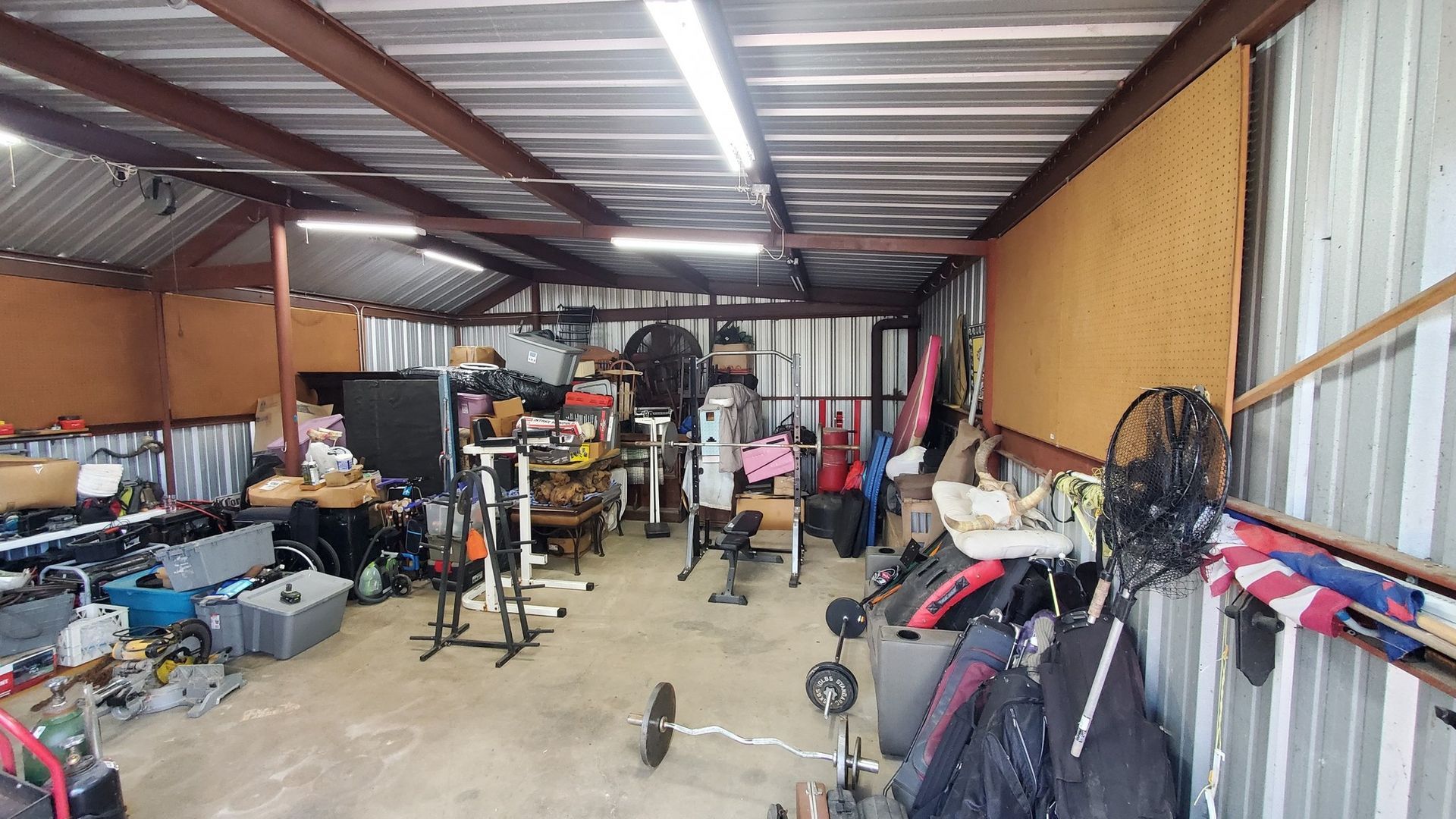 A cluttered garage gym with weightlifting equipment, tools, and storage organized under metal beams and fluorescent lights.