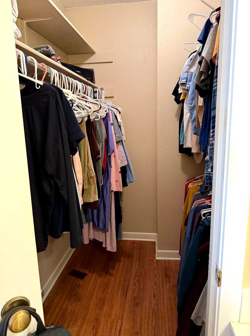 A reach-in walk-in closet with clothes hanging on racks along both side walls, wood-look flooring, and a beige back wall.