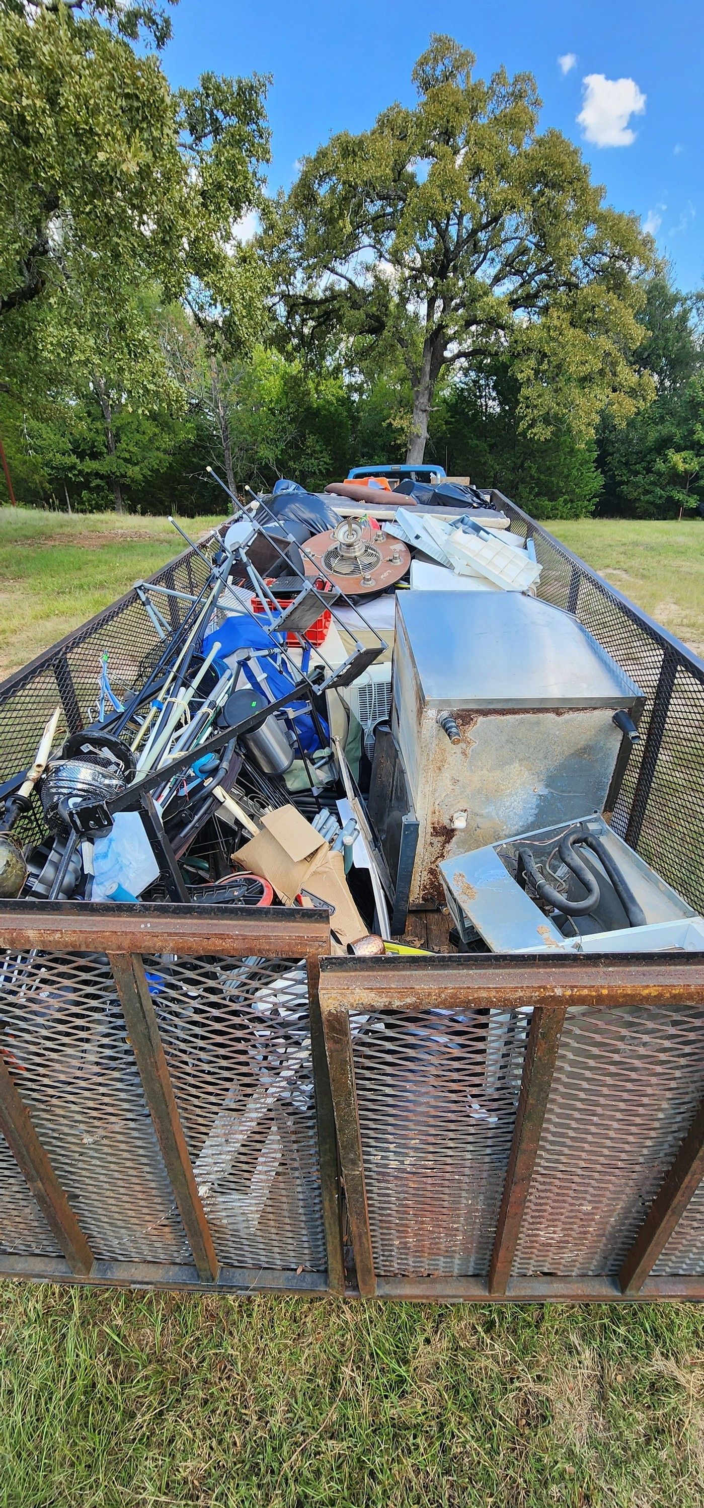 A metal utility trailer filled with various scrap metal, appliances, and debris, parked on a grassy area under a blue sky.