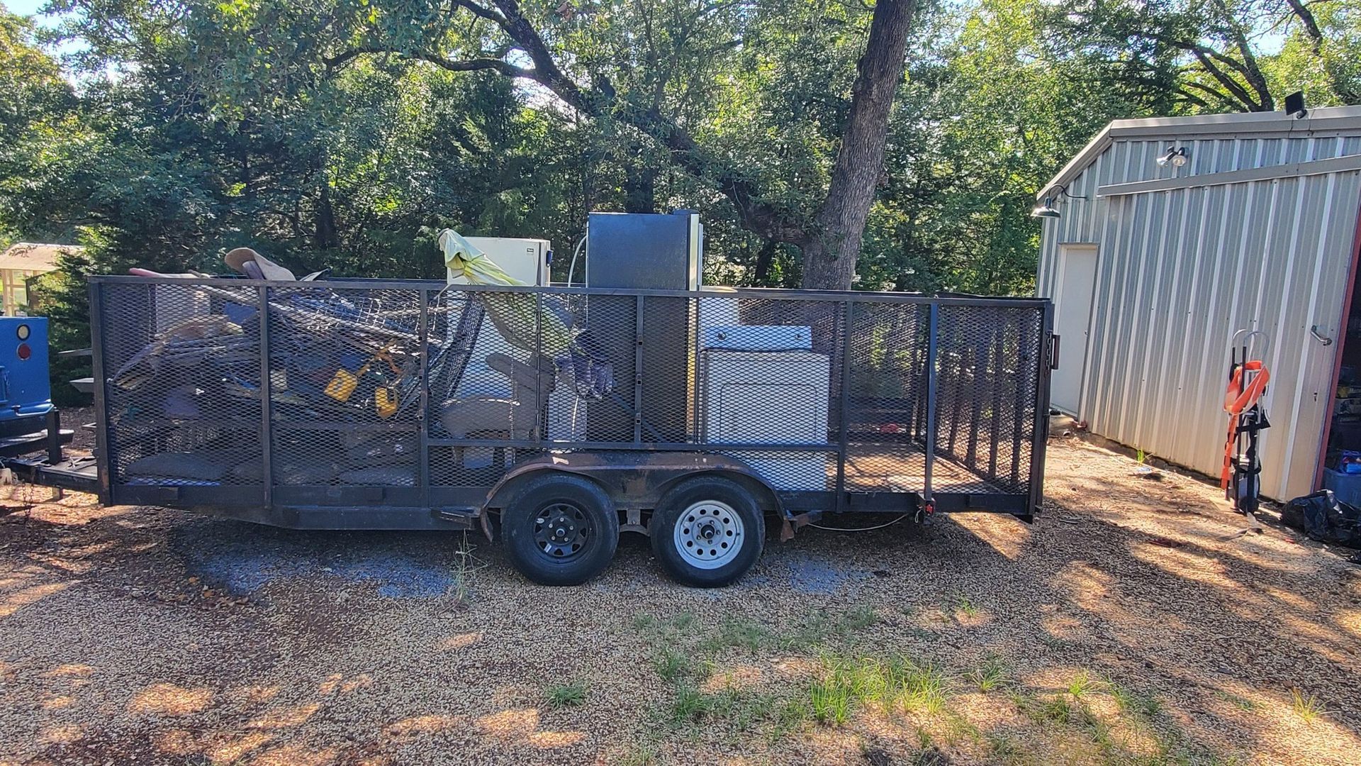 A black utility trailer loaded with various items, including a large dark container and a white box, parked on gravel.