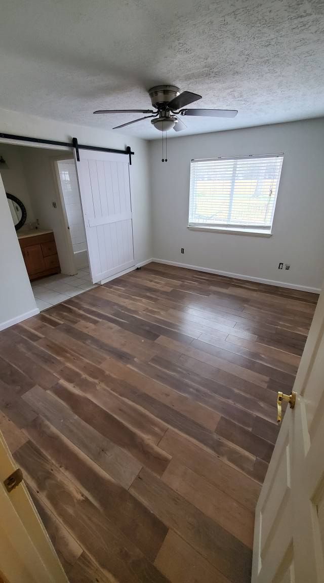 A bright bedroom with wood-look flooring, a ceiling fan, a window with white blinds, and a white sliding barn door.