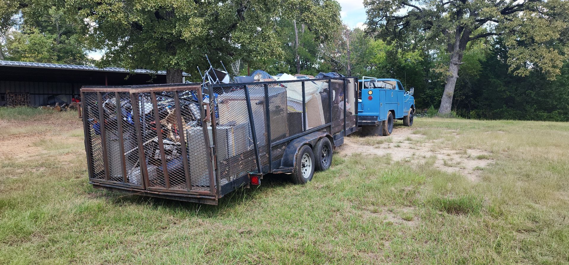 A blue utility truck tows a large metal trailer filled with scrap metal and debris in a grassy field.