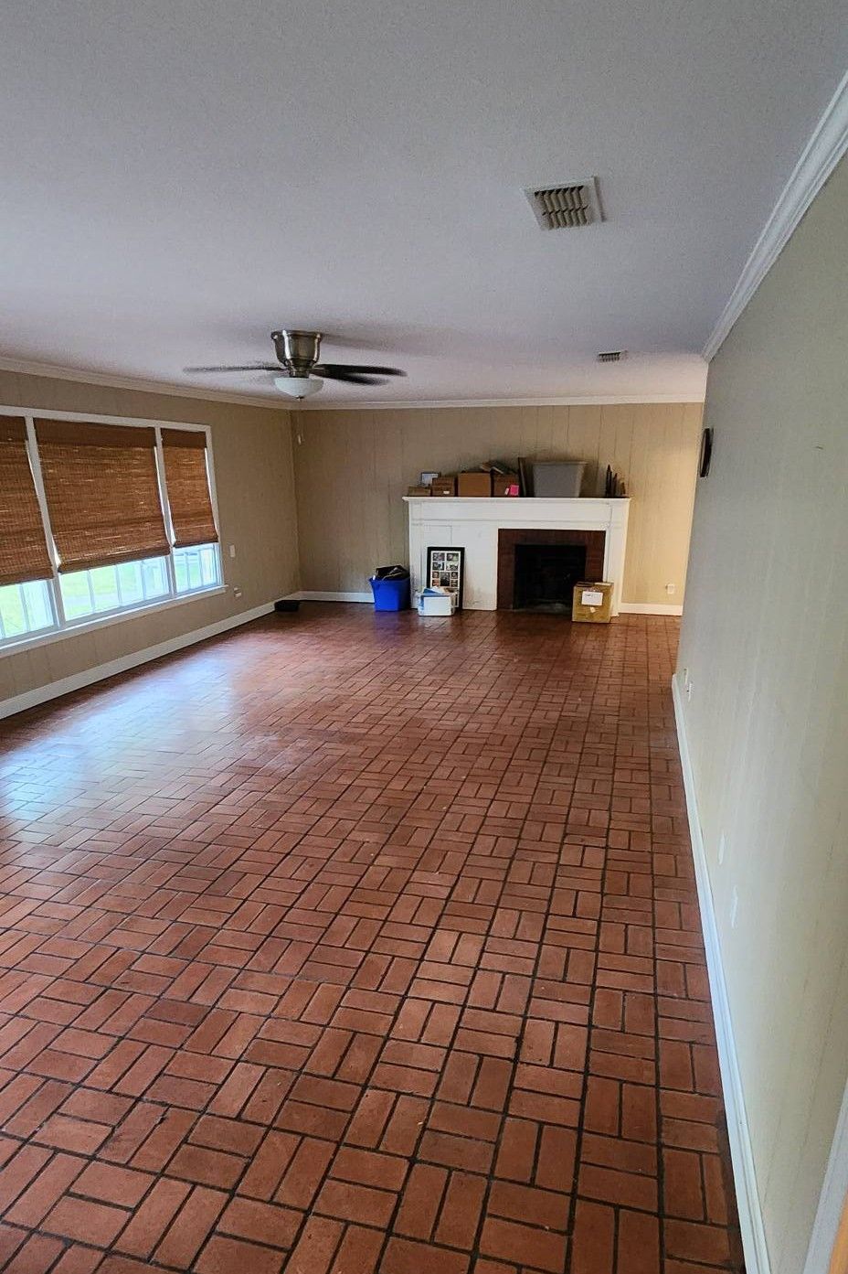 A living room featuring a patterned brick floor, a white fireplace, a ceiling fan, and large windows with bamboo blinds.