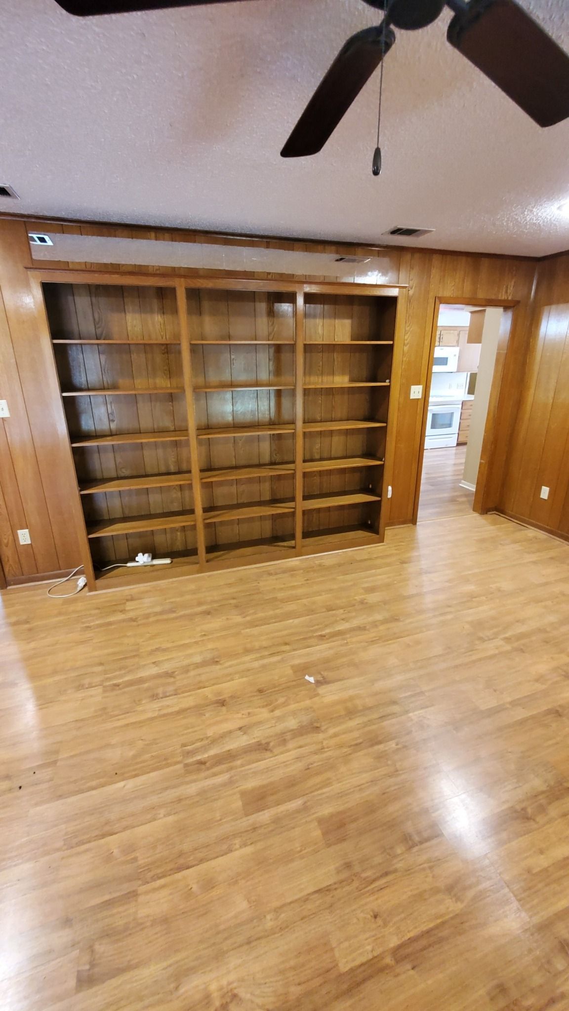 An empty, floor-to-ceiling wooden bookshelf occupies a wall in a room with light wood-look flooring and wood paneling.