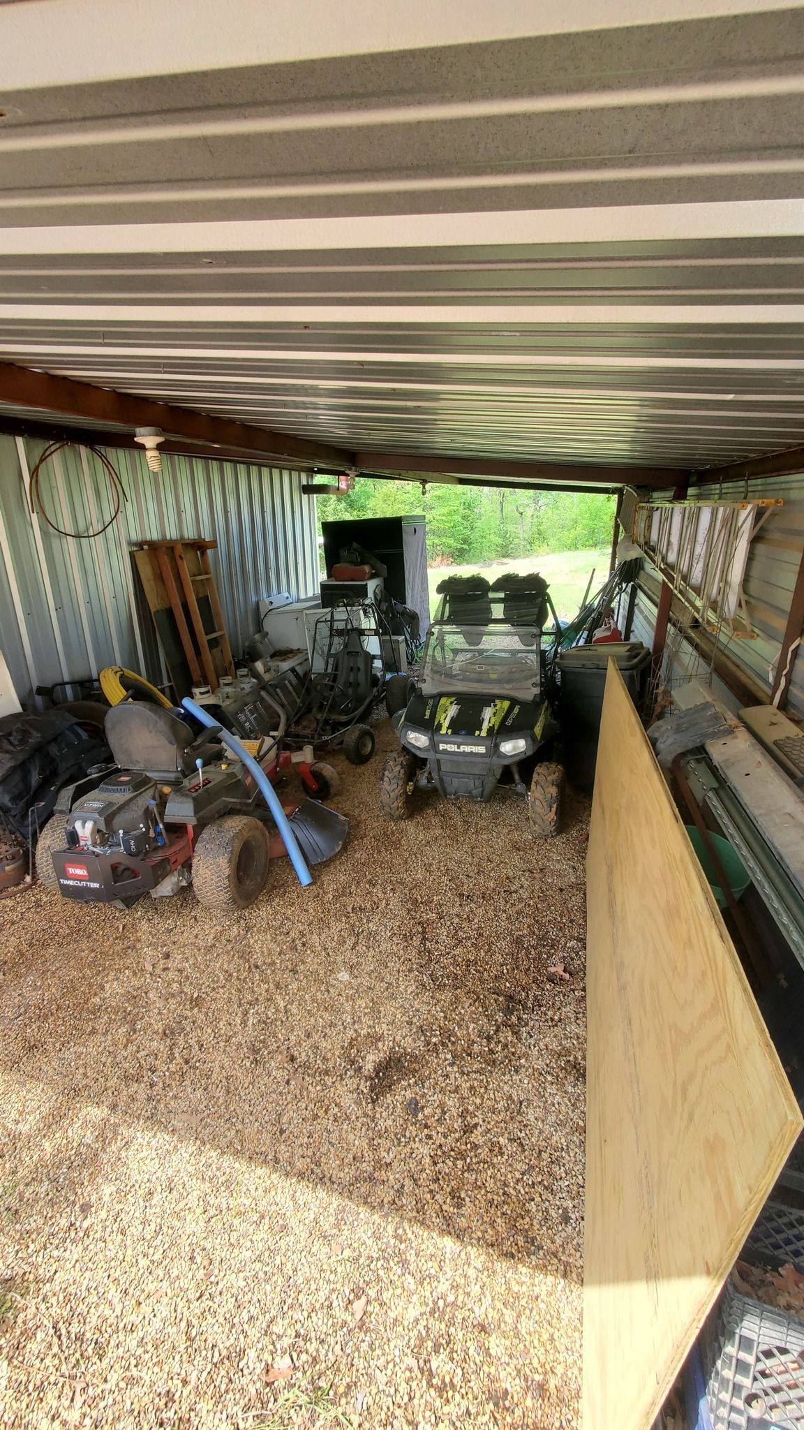 An open-air storage shed filled with a riding lawn mower, an ATV, and other equipment on a wood-chipped floor.
