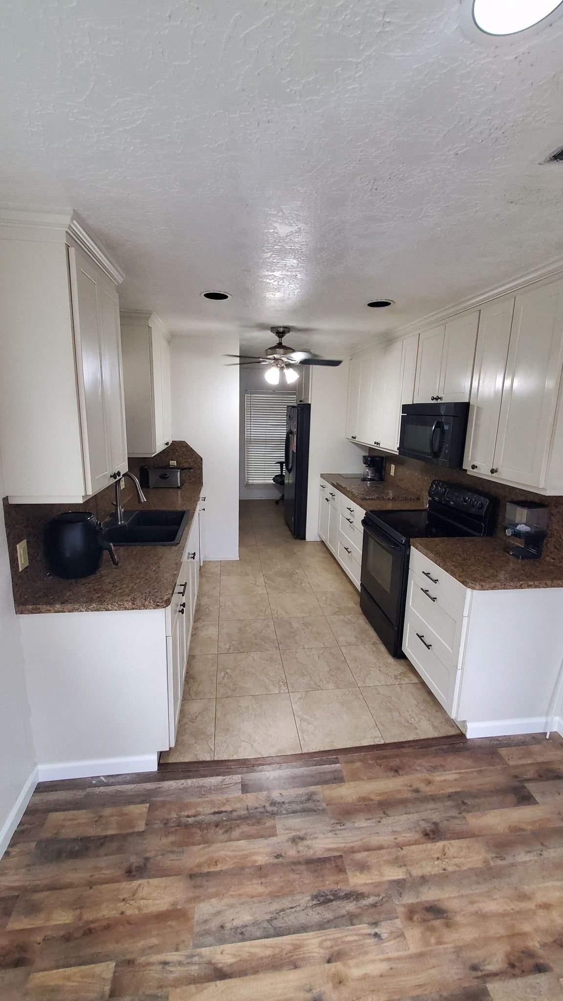A galley-style kitchen with white cabinets, dark countertops, black appliances, and a light tiled floor.