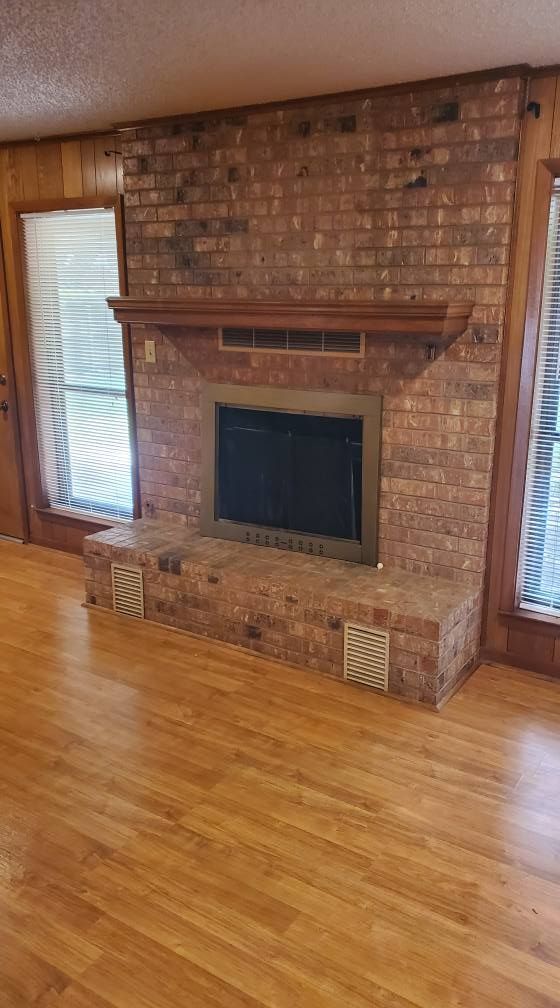 A brick fireplace with a wood mantel and raised hearth sits between two tall windows in a room with wood flooring.