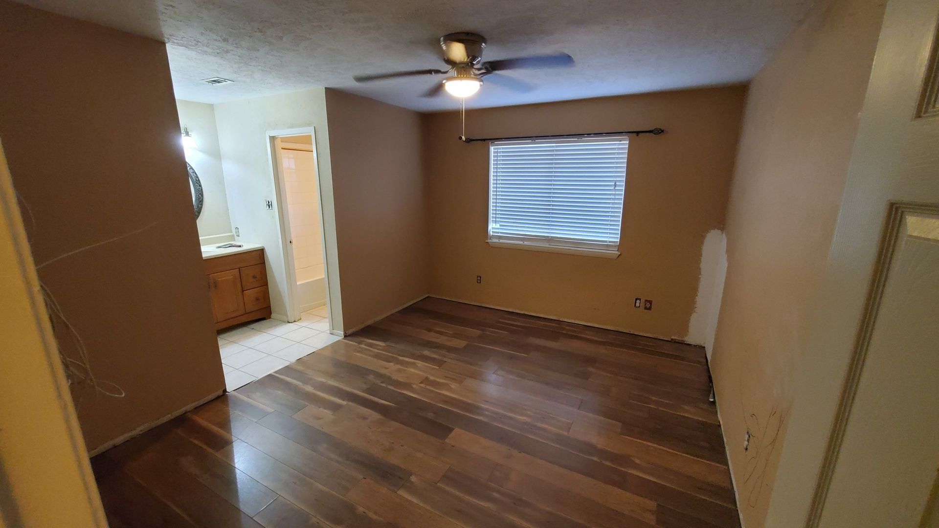 An empty bedroom with tan walls, dark wood-look flooring, a ceiling fan, and a view into an adjoining bathroom.